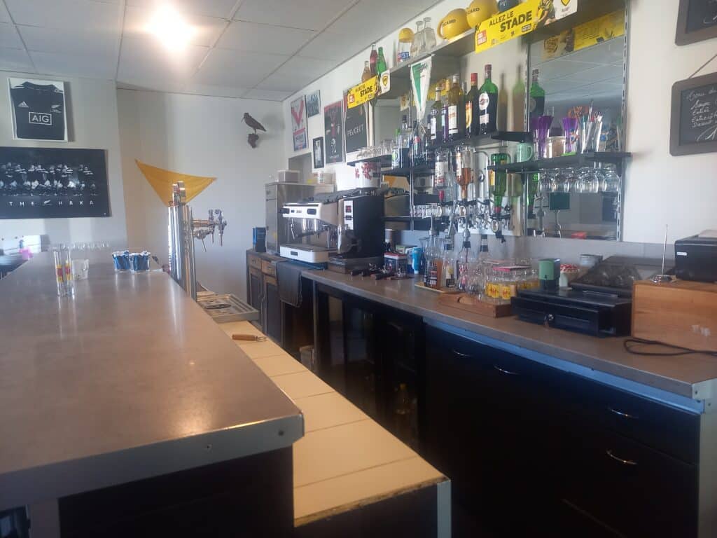 Comptoir de bar professionnel. Arrière-bar avec machine à café, bouteilles (Ricard, Martini), verres. Décor rugby : Maori All Blacks, Haka, Allez le Stade.