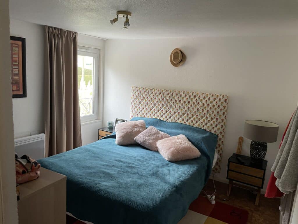 Chambre lumineuse avec grand lit au couvre-lit turquoise, tête de lit fleurie et coussins roses. Tables de chevet, fenêtre, rideaux et chapeau mural.