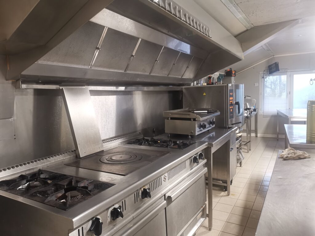 Cuisine professionnelle en inox avec hotte d'extraction, four 'religo!', cuisinière à gaz, plancha et tables de travail en acier. Propre et fonctionnelle.