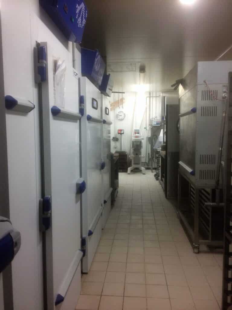 Allée de boulangerie professionnelle avec une rangée de chambres froides blanches, un four, un pétrin et une balance.