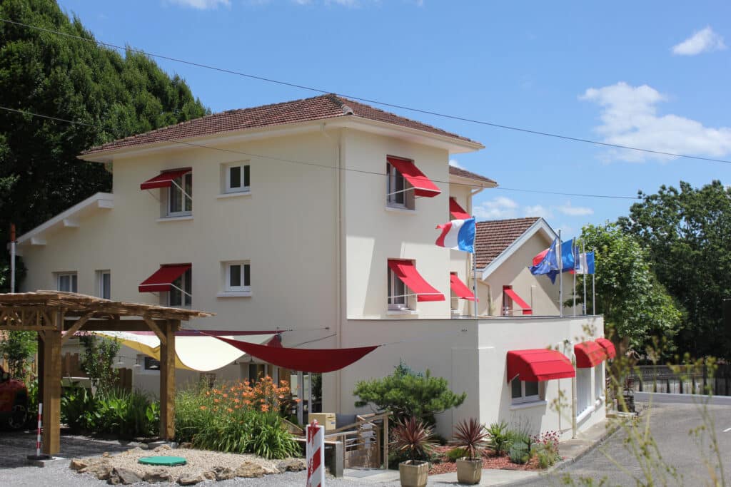 Bâtiment crème avec auvents rouges sous ciel bleu. Pergola en bois, jardin fleuri, et drapeaux français/européen. Hôtel ou résidence ensoleillée.