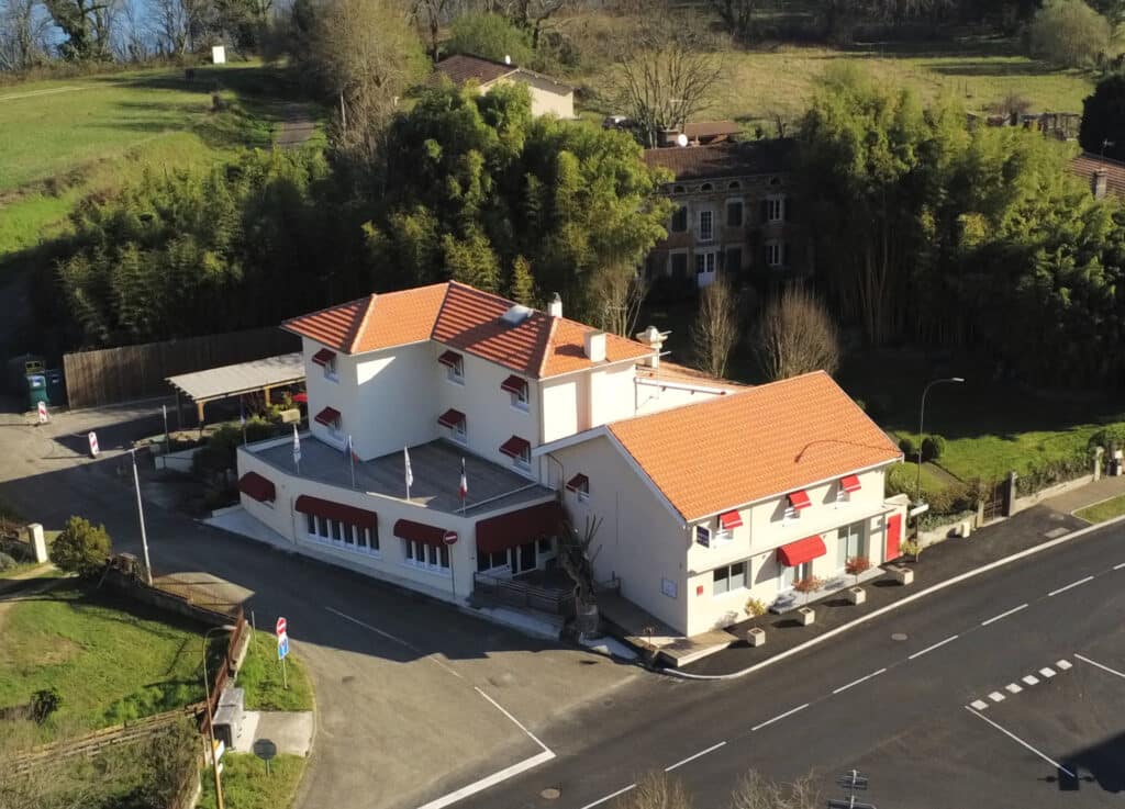 Hôtel avec toits tuiles oranges et auvents rouges, façade blanche, surmonté d'un panneau 'HOTEL', au bord d'une route. Paysage vallonné et verdoyant en arrière-plan.