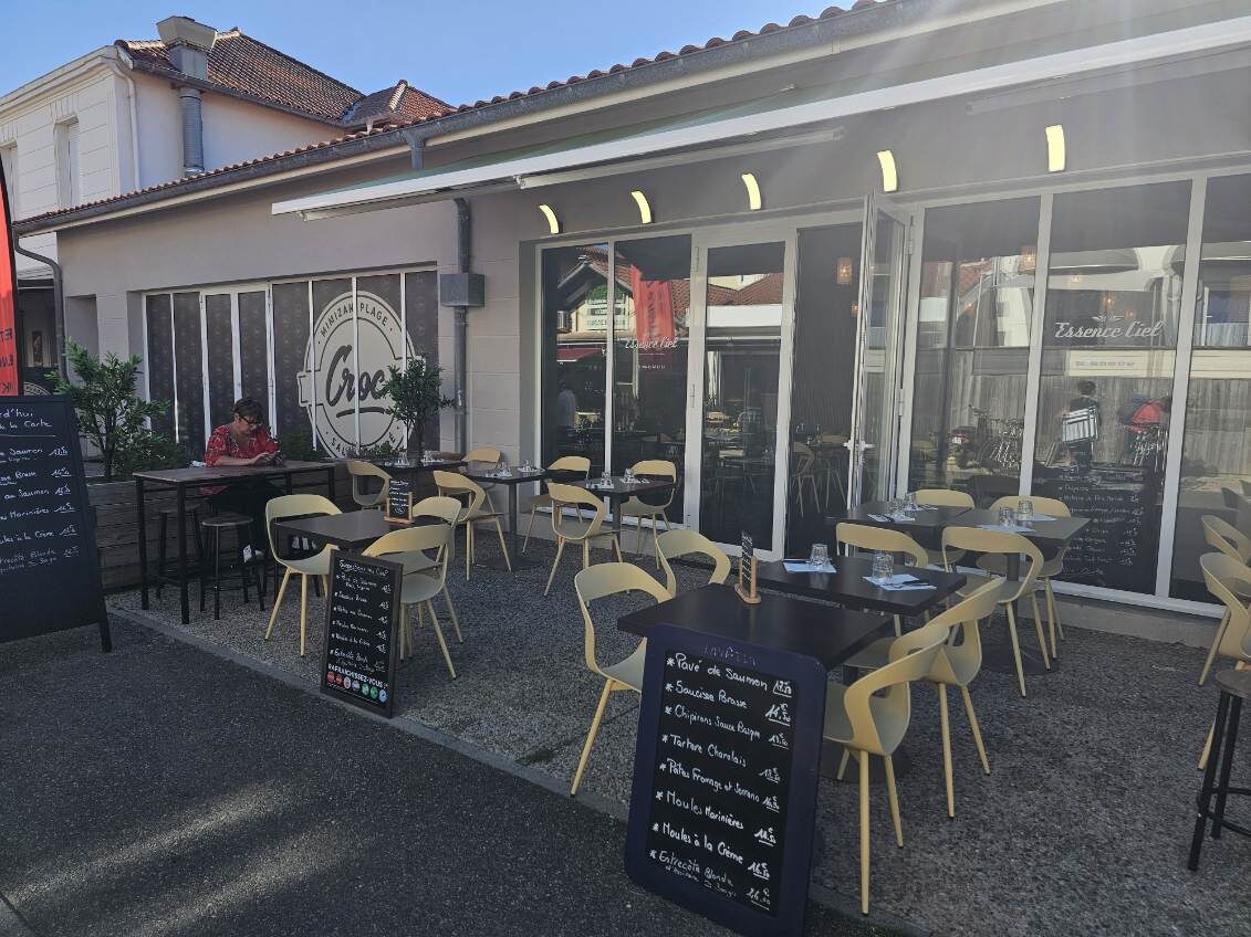 Terrasse extérieure du restaurant Croc Mimizan Plage avec tables et chaises, menus affichés. Une personne consulte son téléphone.