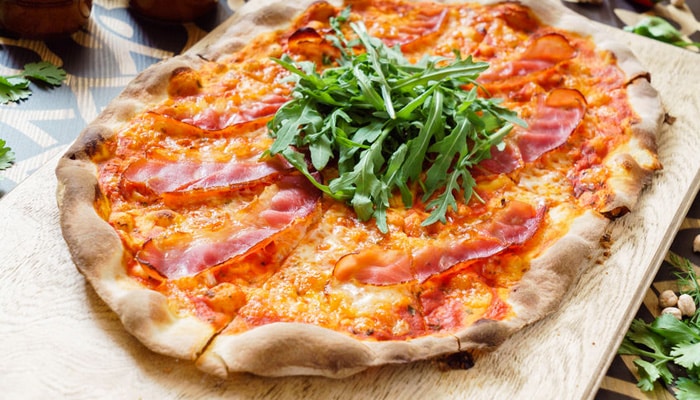 Pizza fine au jambon cru et roquette fraîche sur planche de bois.