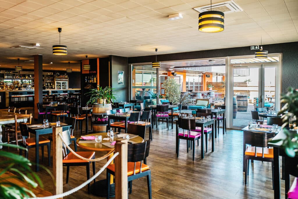 Intérieur moderne du restaurant L'Antre avec tables en bois, chaises colorées et vue sur une terrasse lumineuse.
