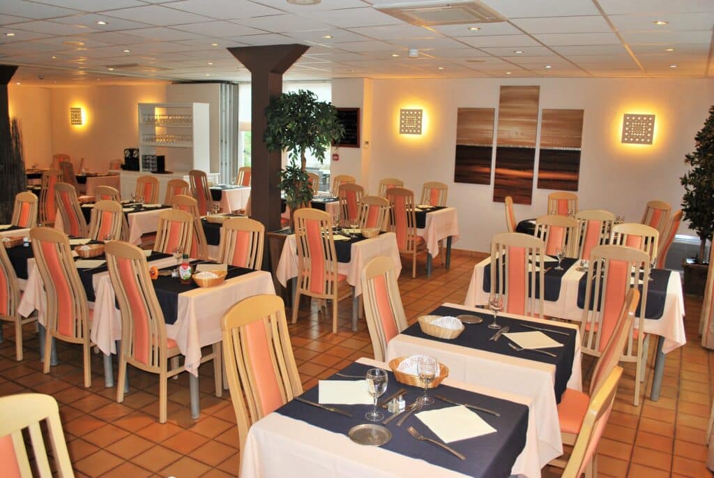 Salle à manger de restaurant élégante : tables dressées, nappes roses, chemins de table bleus, chaises bois clair/orange, décor mural moderne, plantes vertes, éclairage chaleureux.