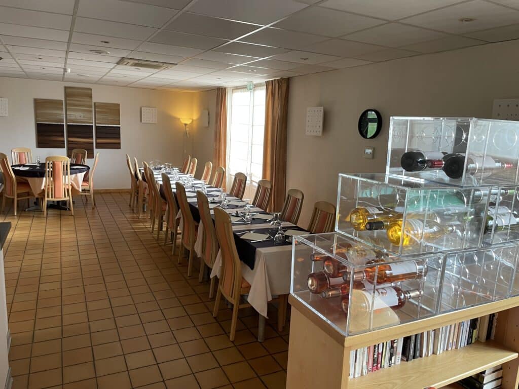 Salle à manger de restaurant avec tables rondes et longue table rectangulaire dressées de nappes, verres. Chaises bois orange. Étagère de vins, livres et sol carrelé.