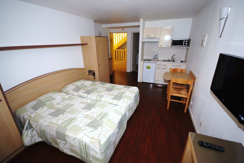 Studio lumineux avec lit double, kitchenette équipée (réfrigérateur, micro-ondes), table repas, TV LG et sol parquet. Escalier jaune en fond.