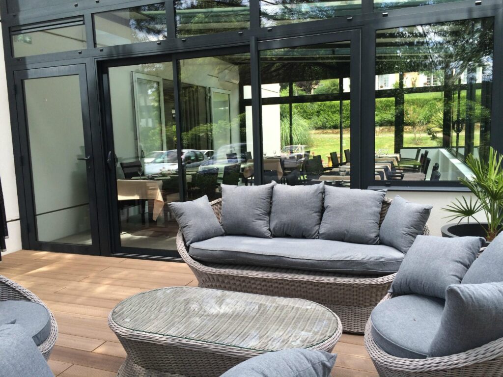 Terrasse bois, mobilier extérieur gris: canapé, fauteuils et table basse. Vue sur un restaurant vitré et un jardin verdoyant.