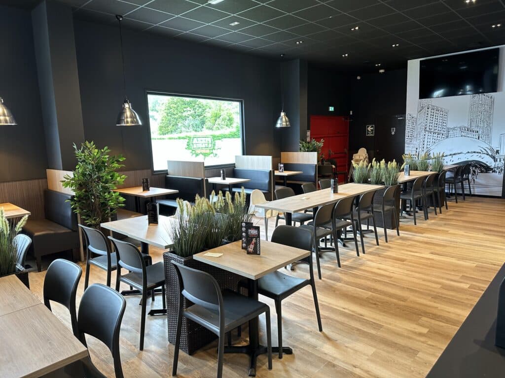 Intérieur restaurant moderne. Murs foncés, mobilier bois/noir, plantes. Vue sur verdure, murale urbaine. Menu 