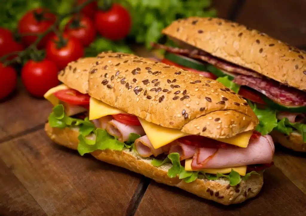Deux appétissants sandwiches garnis de jambon, fromage, tomates et laitue, sur pain aux graines, posés sur une table en bois rustique.