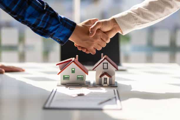 Poignée de main scellant un accord immobilier au-dessus de deux modèles de maisons et d'un contrat sur une table d'agence.