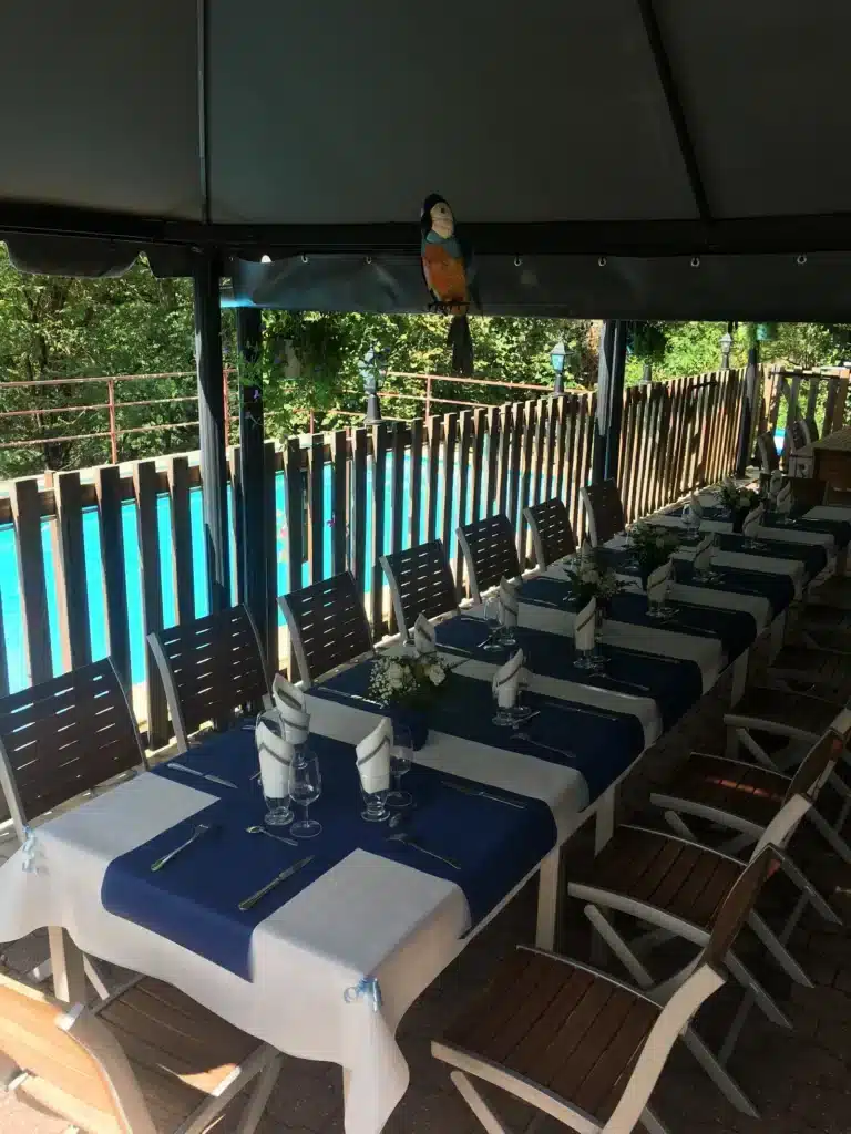 Longue table élégante dressée en extérieur sous un gazebo, avec nappe bleue et décor de perroquet, près d'une piscine.