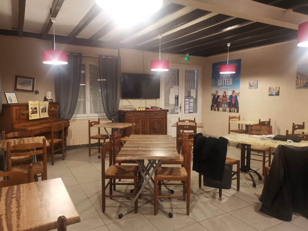 Salle de restaurant rustique : tables et chaises en bois, luminaires rouges. Plafond à poutres sombres. Piano ancien, TV, affiche film PAPI SITTER.