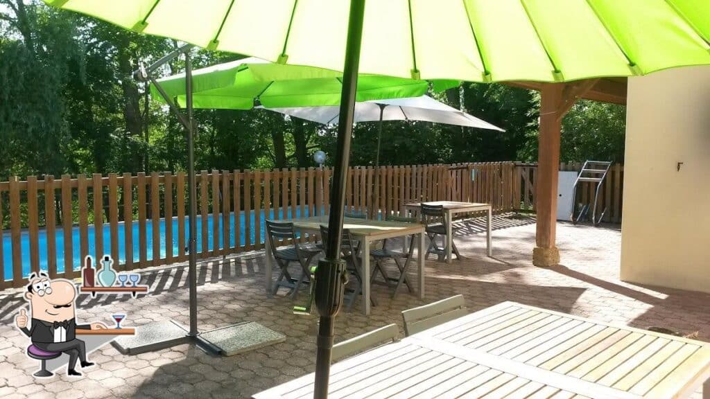 Terrasse ensoleillée avec tables et parasols verts près d'une piscine clôturée, entourée d'arbres verdoyants.