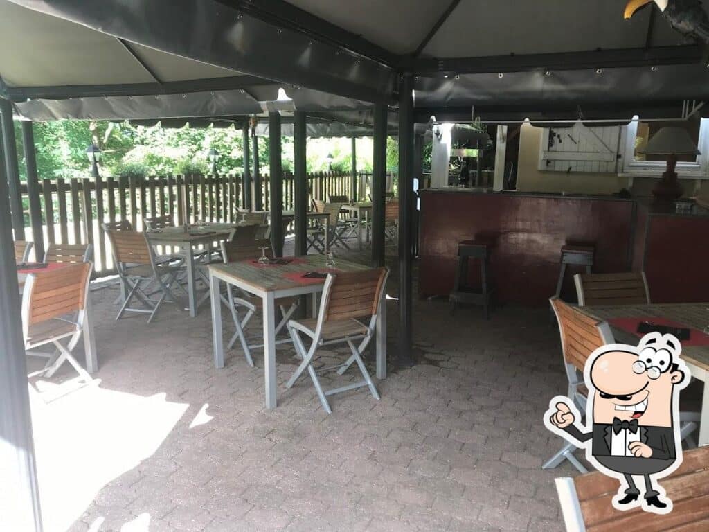 Terrasse de restaurant couverte avec tables et chaises vides, sol pavé, bar et clôture en bois à l'arrière. Cadre extérieur verdoyant.
