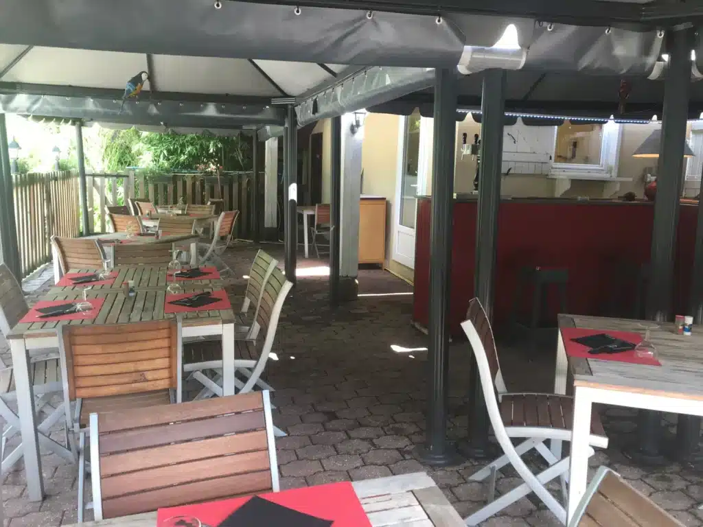 Terrasse de restaurant conviviale avec tables en bois et chaises claires. Un perroquet décoratif est perché sur la bâche. Bar rouge et verdure en fond.