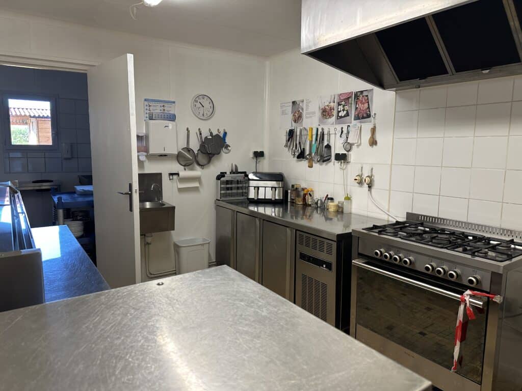 Cuisine professionnelle en inox avec four Bartscher, plaque gaz, évier. Plan de travail, ustensiles. Poster hygiène 'LAVAGE MAINS', horloge 7h50.