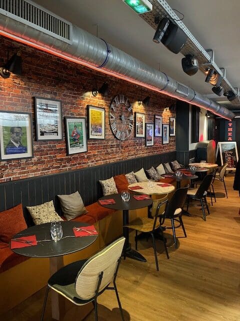 Restaurant-bar moderne: mur de briques, affiches, horloge, banquettes rouges, tables préparées, éclairage industriel. Panneau 'Comedy Club'.
