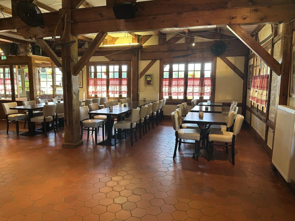 Intérieur de restaurant rustique avec poutres en bois, tables et chaises claires. Sol en carreaux hexagonaux terre cuite. Fenêtres avec rideaux à carreaux rouges.