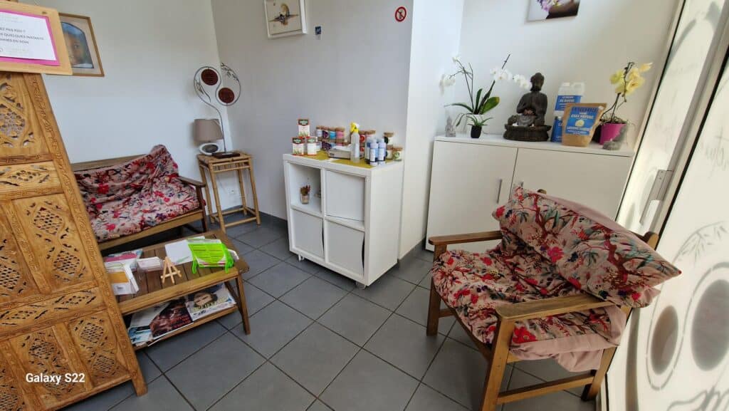 Salle d'attente accueillante avec mobilier en bois sculpté, coussins floraux, statue de Bouddha, orchidées. Texte: 
