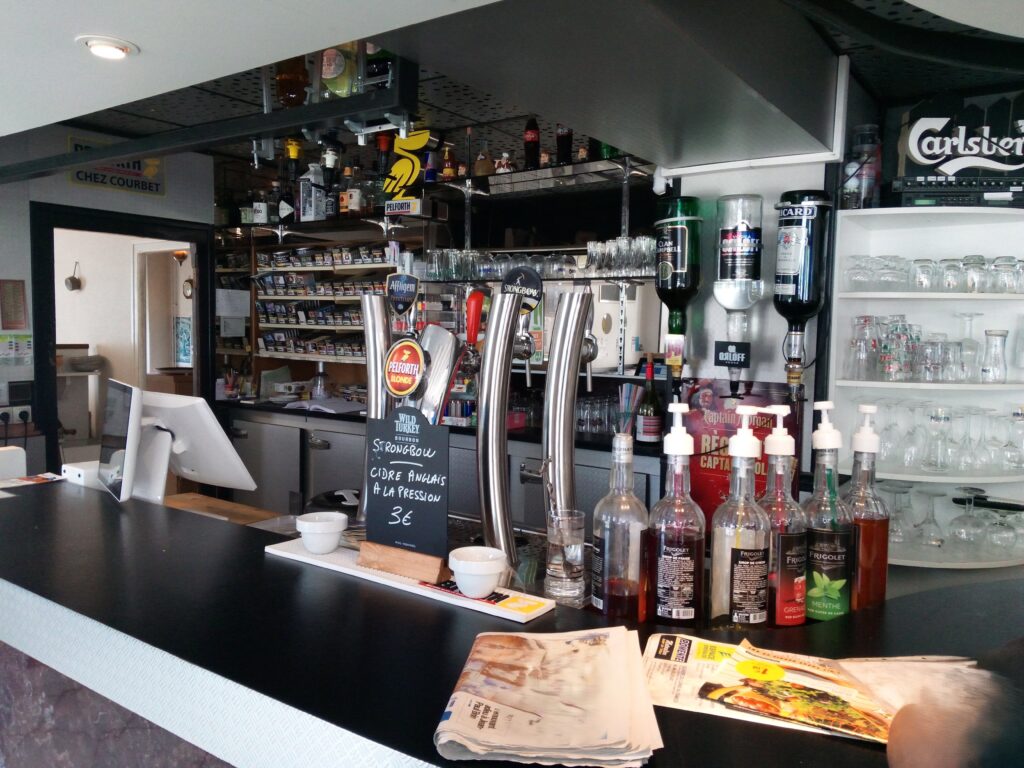 Bar moderne Chez Courbet: comptoir noir, tireuses Pelforth & Strongbow (cidre pression 3€), distributeurs d'alcool, étagères de verres et cigarettes.