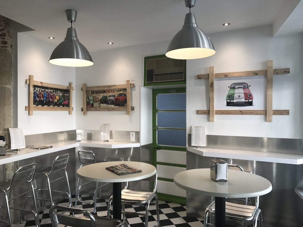 Café italien rétro : luminaires industriels, carrelage damier noir et blanc, affiches 'ITALIAN DREAM', Vespa et Fiat 500 'Viva Italia', chaises chromées.