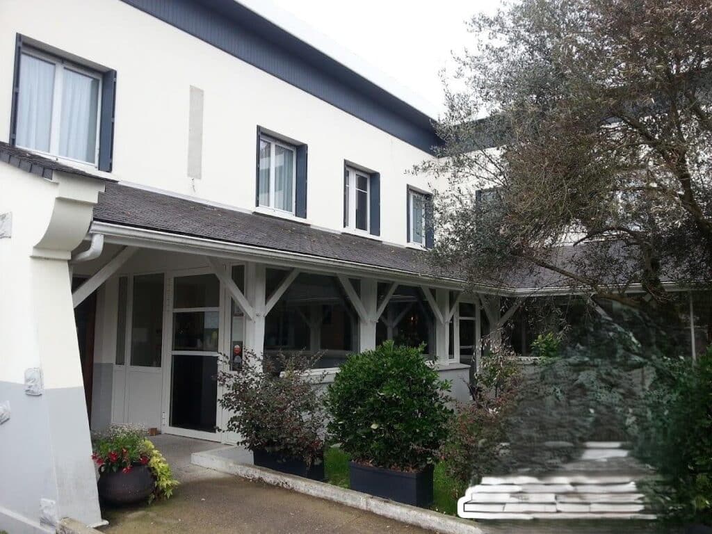 Façade blanche d'un hôtel avec entrée couverte et véranda. Fenêtres à volets foncés, plantes en pot et grand arbre devant.