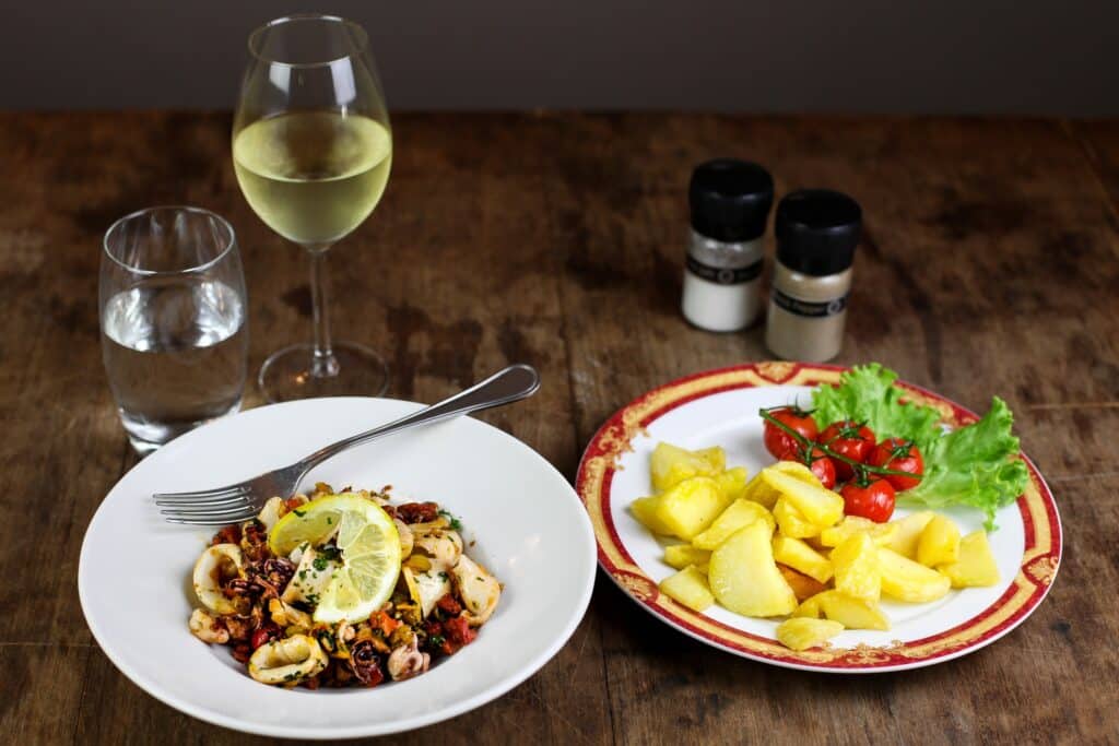 Repas méditerranéen sur table en bois: calamars citronnés, pommes de terre, tomates cerises, salade, vin blanc, eau. Salière et poivrier 'Black Pepper'.