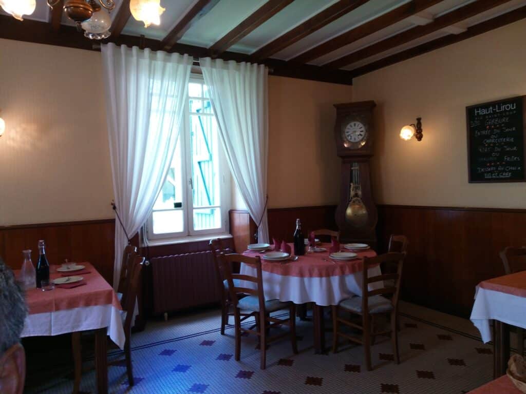 Intérieur de restaurant avec tables nappées, rideaux blancs. Horloge comtoise et menu 'Haut-Lirou' lisible: Garbure, Plat du Jour, Grillade, Vin.