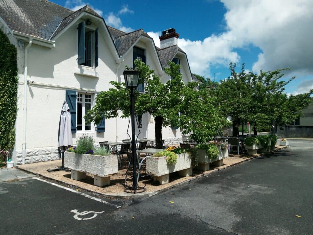 Bâtiment blanc avec volets bleus, toit gris, terrasse extérieure ombragée de verdure. Tables, chaises, jardinières fleuries et place PMR au sol.