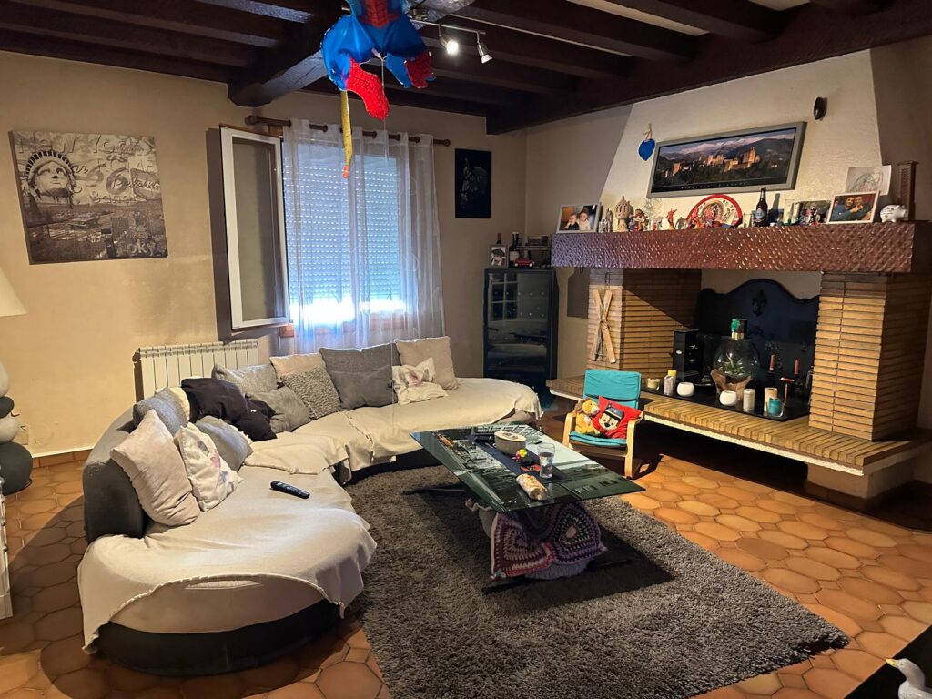 Salon chaleureux avec grand canapé beige en U, cheminée en brique décorée de photos et bibelots. Tapis moelleux gris. Spiderman pend du plafond.