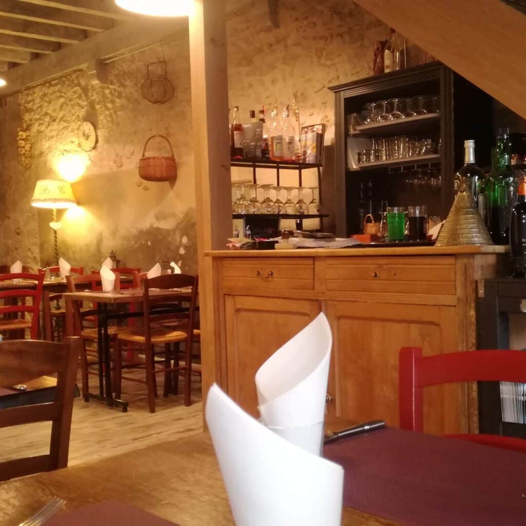 Ambiance chaleureuse de restaurant rustique avec mur en pierre, comptoir en bois, tables dressées avec serviettes en papier pliées et lampes tamisées.