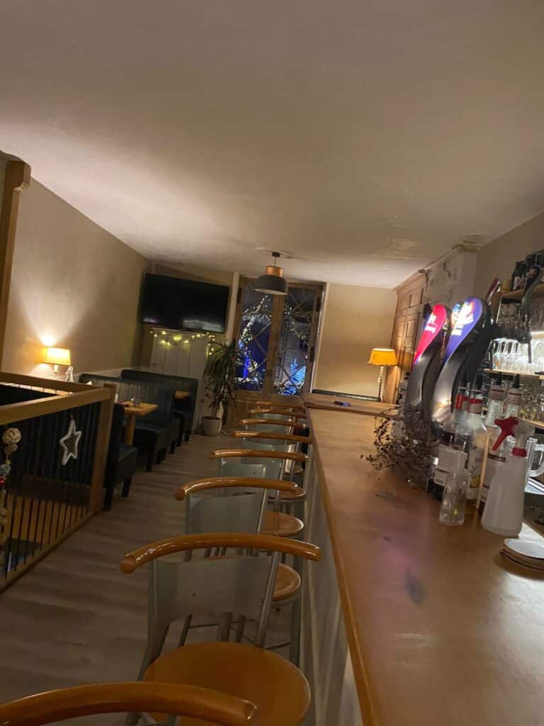 Intérieur de bar ou café accueillant, éclairage tamisé, tabourets bois/métal alignés devant un comptoir en bois, tireuses et bouteilles visibles.