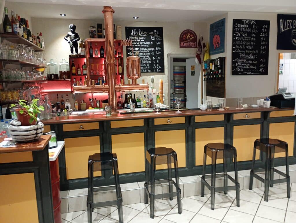 Bar intérieur avec tireuses à bière en cuivre, comptoir en bois, tabourets noirs et menus 'Nos Bières' et 'Tapas' affichés.