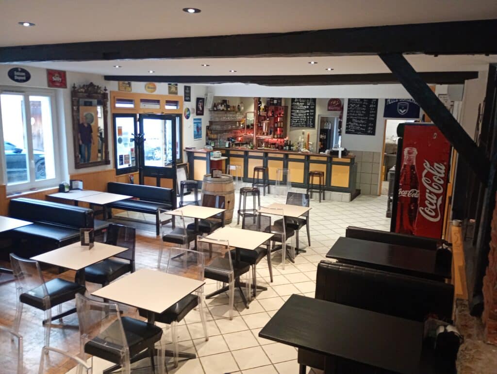 Intérieur de café ou pub avec poutres sombres, tables claires et chaises transparentes. Panneau Coca-Cola rouge visible.