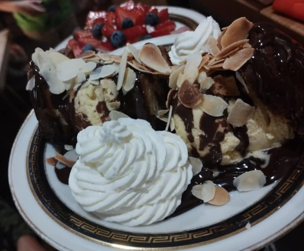 Gâteau moelleux nappé de chocolat, glace vanille, amandes effilées, chantilly, et fruits rouges en arrière-plan.