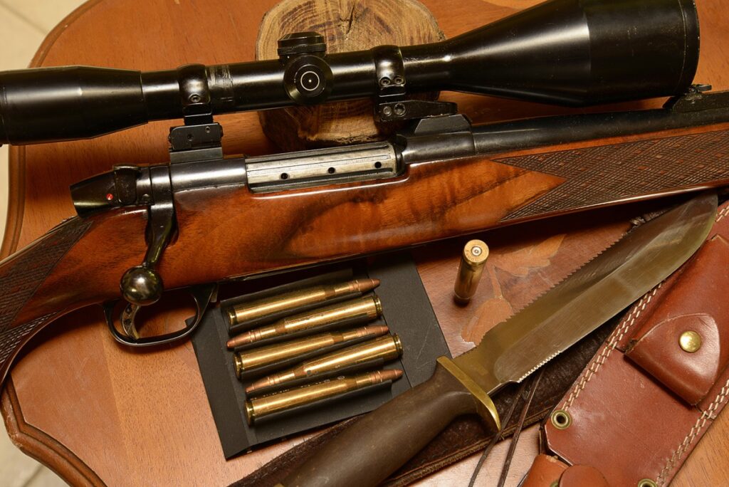 Carabine de chasse à verrou avec lunette, munitions et grand couteau de chasse à lame dentelée sur fond boisé.
