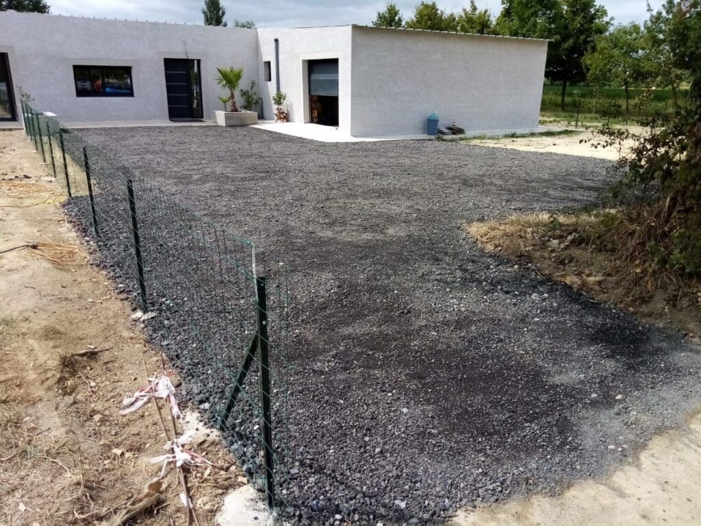 Allée en gravier noir fraîchement posé devant une maison moderne blanche, délimitée par une clôture grillagée verte.