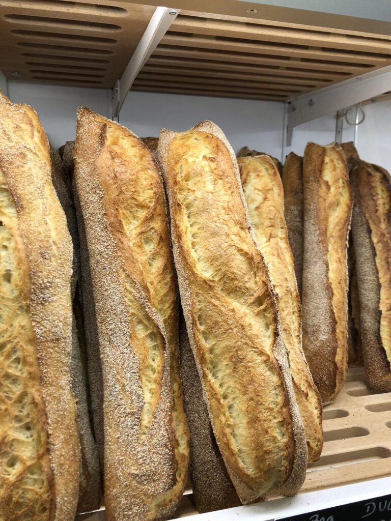 Baguettes françaises dorées et croustillantes, saupoudrées de farine, alignées sur des étagères en bois dans une boulangerie. Texte visible : DUO.