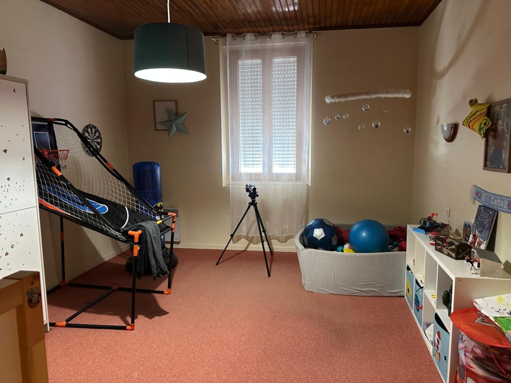Chambre d'enfant ou salle de jeux avec tapis rouge, panier de basket, divers ballons, trépied photo et étagères de jouets. On aperçoit l'inscription 