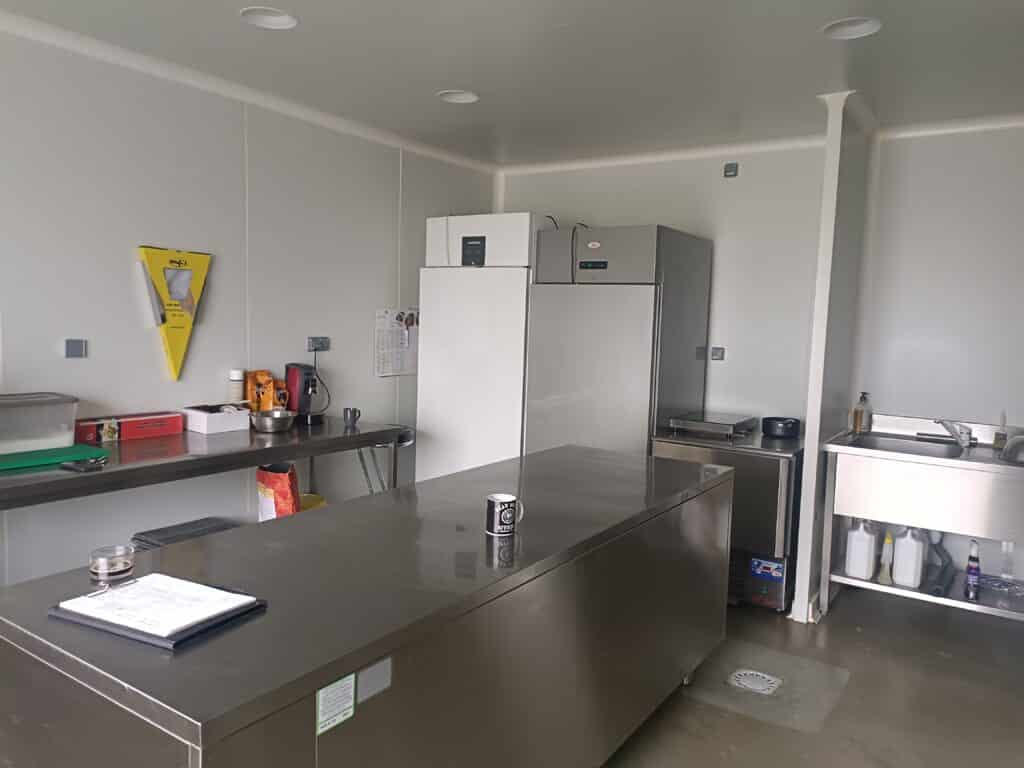 Cuisine professionnelle ou vestiaire avec plan de travail en inox, deux réfrigérateurs/congélateurs, évier, et distributeur de sacs 