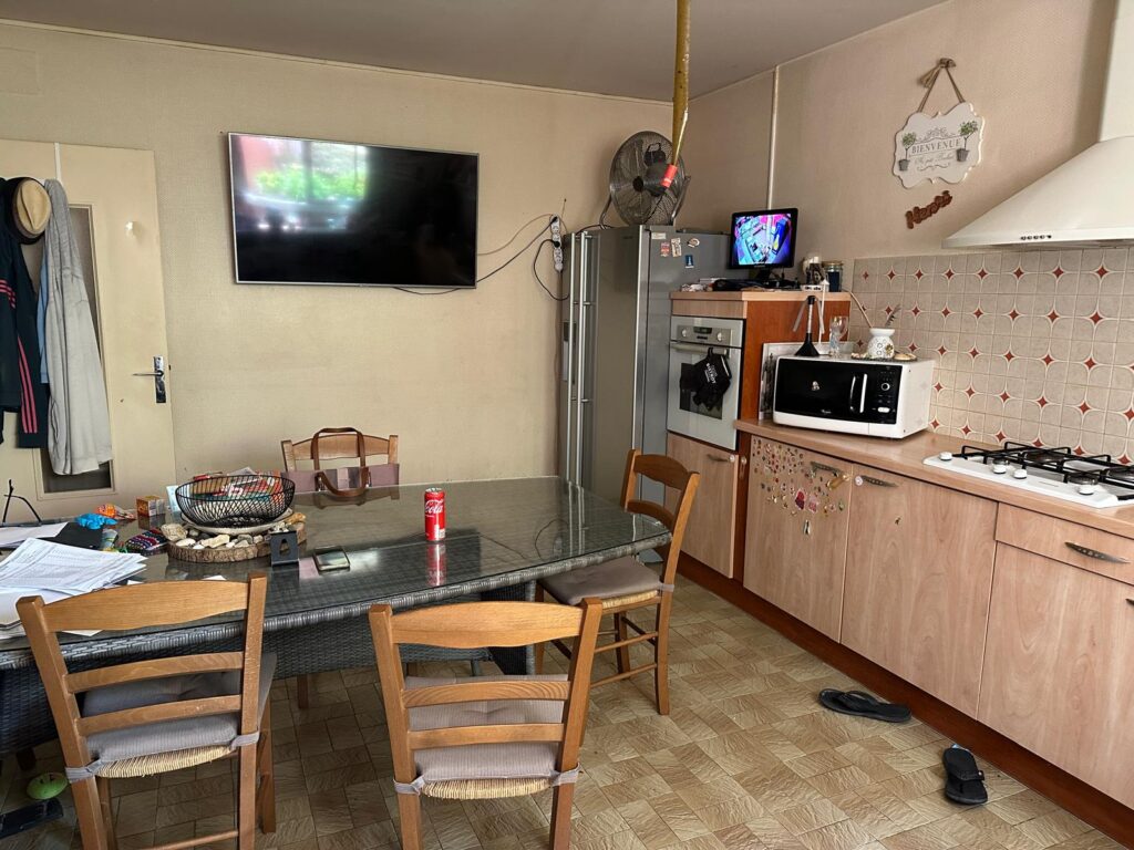 Cuisine salle à manger avec table en verre, chaises en bois, TV murale et électroménagers, panneau 'BIENVENUE' visible.