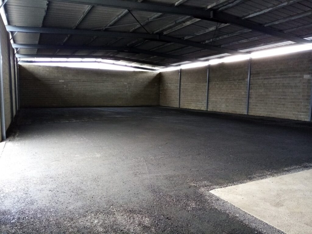 Intérieur d'un hangar vide avec toit métallique, murs en parpaings et sol fraîchement asphalté contrastant avec une dalle de béton.