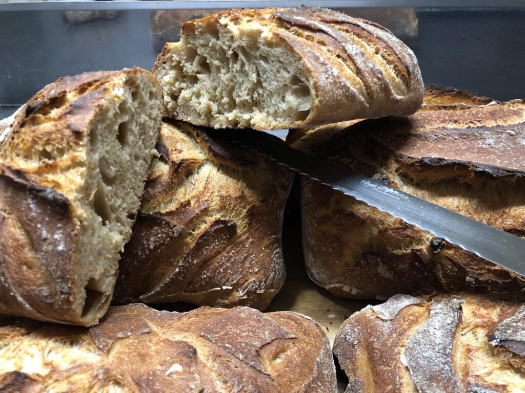 Gros plan sur une pile de pains au levain rustiques, dorés et croustillants, dont deux sont coupés pour montrer la mie alvéolée. Un couteau à pain est visible.