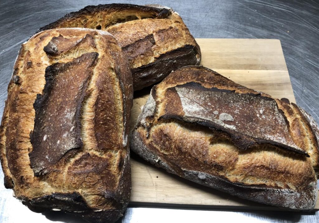 Trois pains au levain rustiques, bien dorés et fortement grillés avec des ouvertures marquées, posés sur une planche à découper en bois.