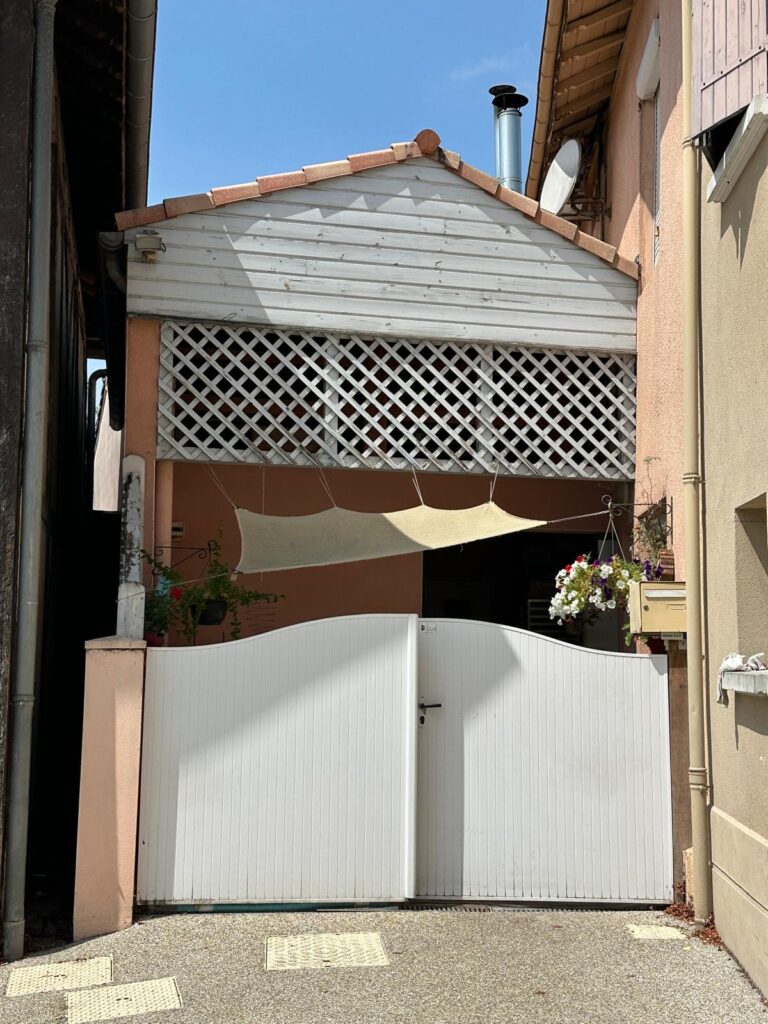 Portail blanc ouvrant sur une cour avec façade en bois clair, treillis décoratif et fleurs suspendues sous un ciel bleu.