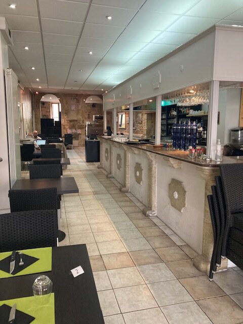 Intérieur de restaurant ou café long et étroit avec un long comptoir de bar décoratif à droite et des tables noires et chaises en osier à gauche. Une table porte le numéro '2'.