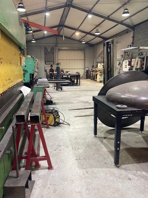 Vaste atelier de fabrication métallique. Grande presse plieuse industrielle, pièces sur tréteaux et table de soudage avec pièce bombée. Un soudeur travaille.