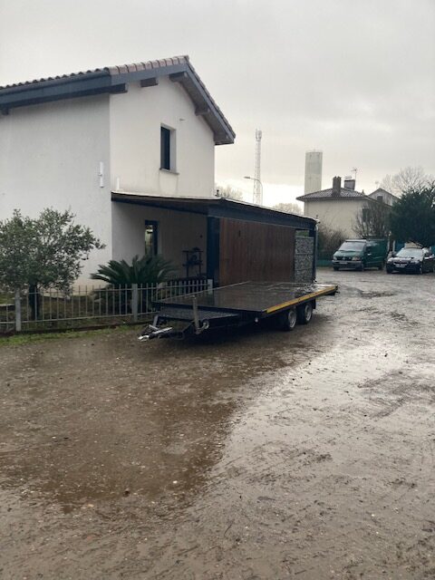 Remorque plateau sur sol boueux et humide devant une maison blanche et un carport. Au loin : véhicules, tour de communication et château d'eau.